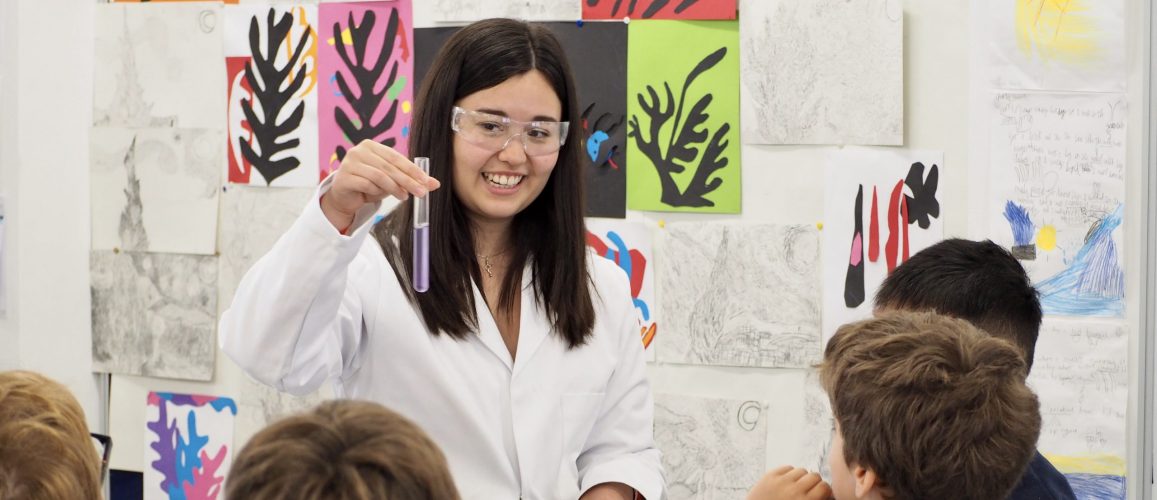 A female scientist in a lab coat holding a vial of liquid showing it to some pupils.